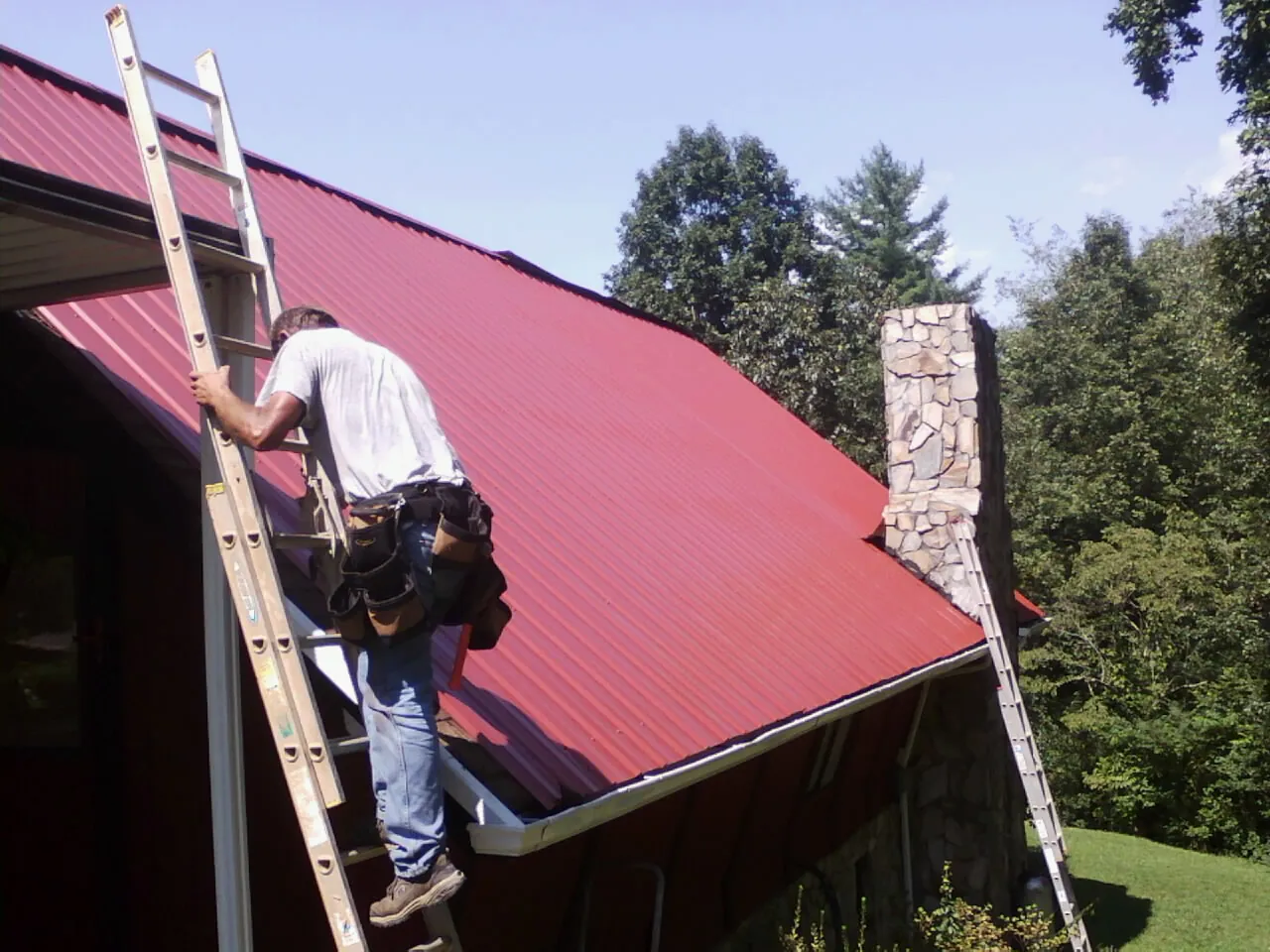 Roofer on a ladder beside a red metal roof and stone chimney — Soffit & Fascia Repair by Roofing Brothers in Medora